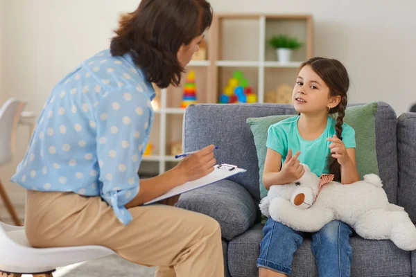 Счастливая маленькая девочка рассказывает историю детскому психологу во время сеанса терапии в уютном современном офисе — стоковое фото