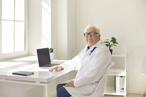 Pozytywny starszy lekarz w mundurze siedzi z laptopem w gabinecie medycznym i patrzy w kamerę — Zdjęcie stockowe