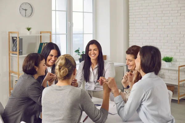 Team of happy young business women sitting around office table and holding hands — Stock Photo, Image