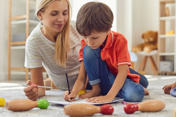 Happy young mother helping her little son to draw on paper on floor at home