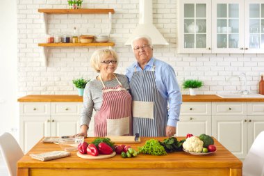 Mutfakta önlüklü yaşlı çift yemek pişirme ürünlerinin yanında kameraya bakıyor..