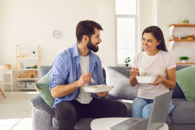 Evli bir çift ellerinde yemek tepsisi ve kanepede oturup yemek yerken konuşuyor..