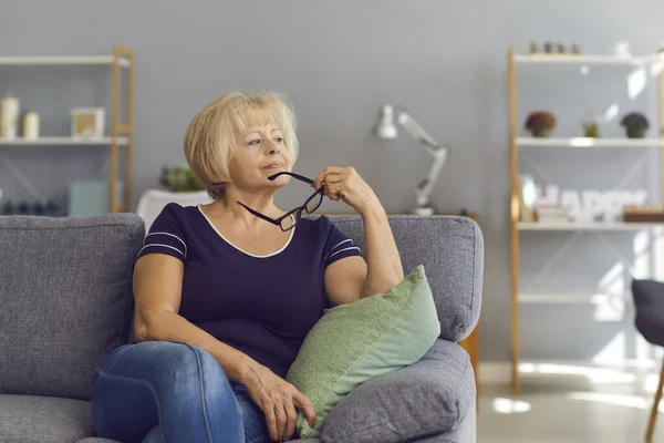 Relaxed mature woman sitting on sofa, holding glasses, looking away and thinking