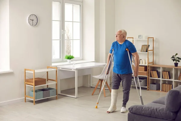 Starší muž se zlomenou nohou v sádře chodí po obýváku a používá berle. — Stock fotografie