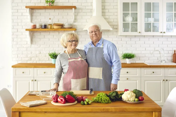 Senior couple standing in aprons in the kitchen near cooking products and looking at camera.