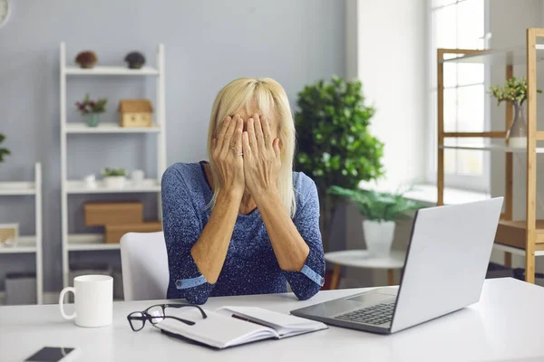 Upprörd äldre kvinna sitter vid skrivbordet med bärbar dator och täcker ansiktet oförmögen att hantera problem — Stockfoto