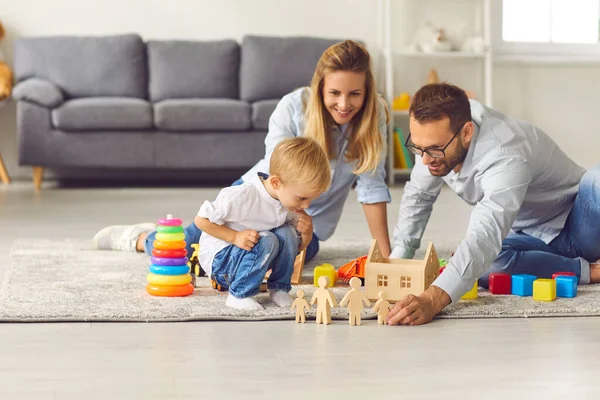 Young parents play with their son wooden figurines sitting on the floor among various toys.