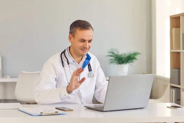 Uśmiechnięty lekarz e-zdrowia macha ręką na laptopie, witając pacjenta podczas rozmowy wideo — Zdjęcie stockowe