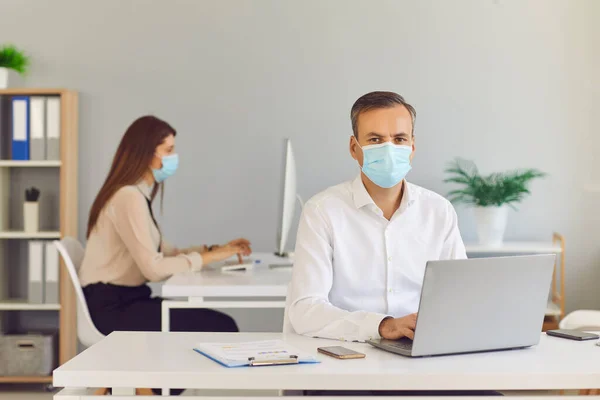 Serious young man in medical face mask working in the company office with his colleague