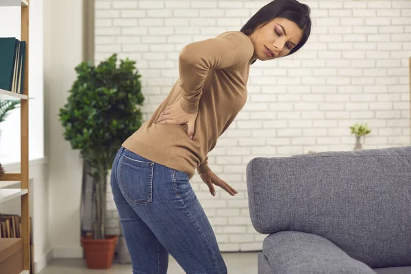 Young woman stands up from sofa and feels sudden intense pain in her lower back — Stock Photo, Image