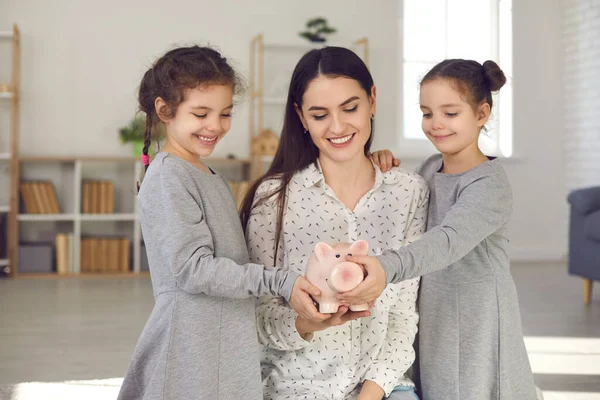 Gelukkig jong moeder en haar twee schattig kleine dochters houden spaarvarken bank en glimlachen — Stockfoto