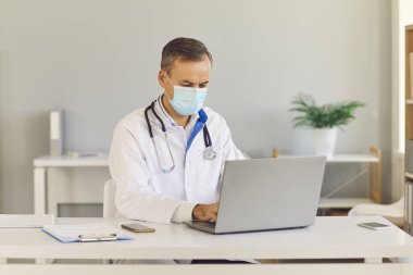 Orta yaşlı, koruyucu maske takan ciddi bir doktor. Oturuyor ve hastaya danışmanlık yapıyor.