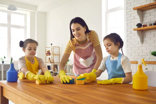 Щасливі маленькі дочки допомагають мамі помити стіл губками та миючим засобом — стокове фото