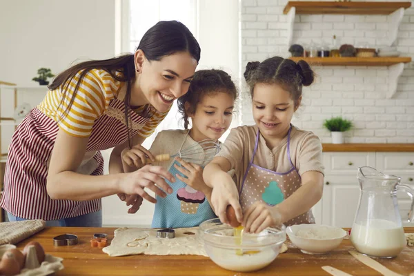 Happy young mother and cute helpful little daughters making dough for cookies together