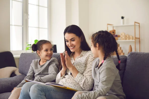 Opiekuńcza uśmiechnięta mama czyta książkę z dwiema córkami siedzącymi na kanapie w domu. — Zdjęcie stockowe