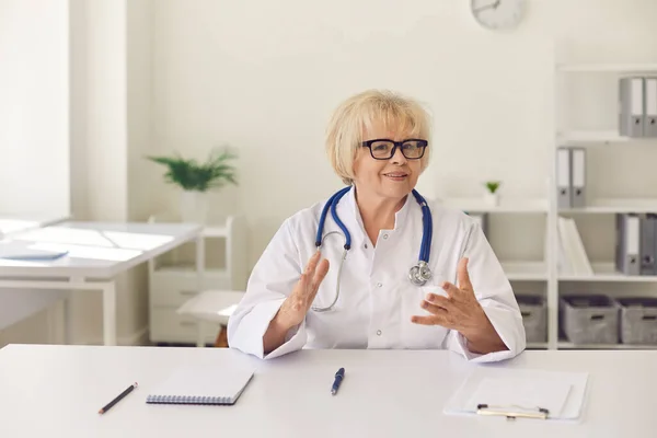 Sourire confiant femme âgée médecin assis et regardant la caméra pendant le vidéocall en ligne — Photo