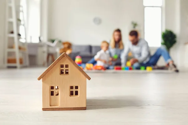 Close-up van een klein houten huis op de achtergrond van een jong gezin dat droomt van hun huis. — Stockfoto