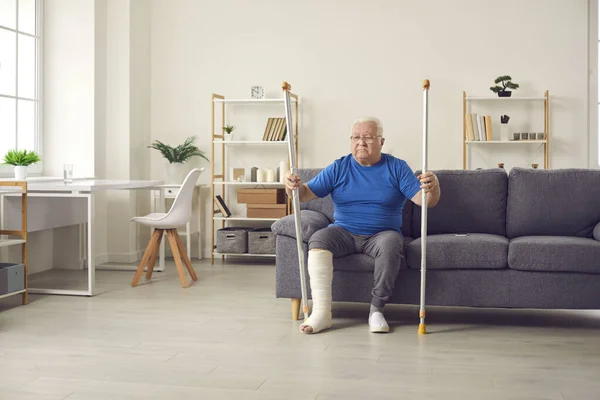 Старший чоловік з розбитою ногою в кастингу тримає милиці і хоче встати з дивана — стокове фото