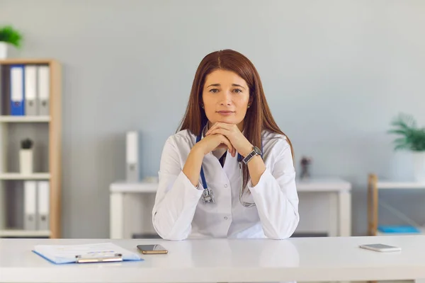 Confident young female doctor sitting at desk, looking at camera and listening to you — Stock Photo, Image