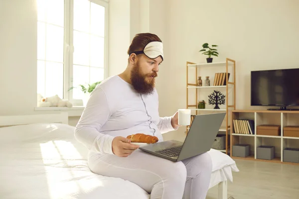 Забавный человек в белой пижаме сидит с кофе и круассаном в постели и смотрит на ноутбук — стоковое фото
