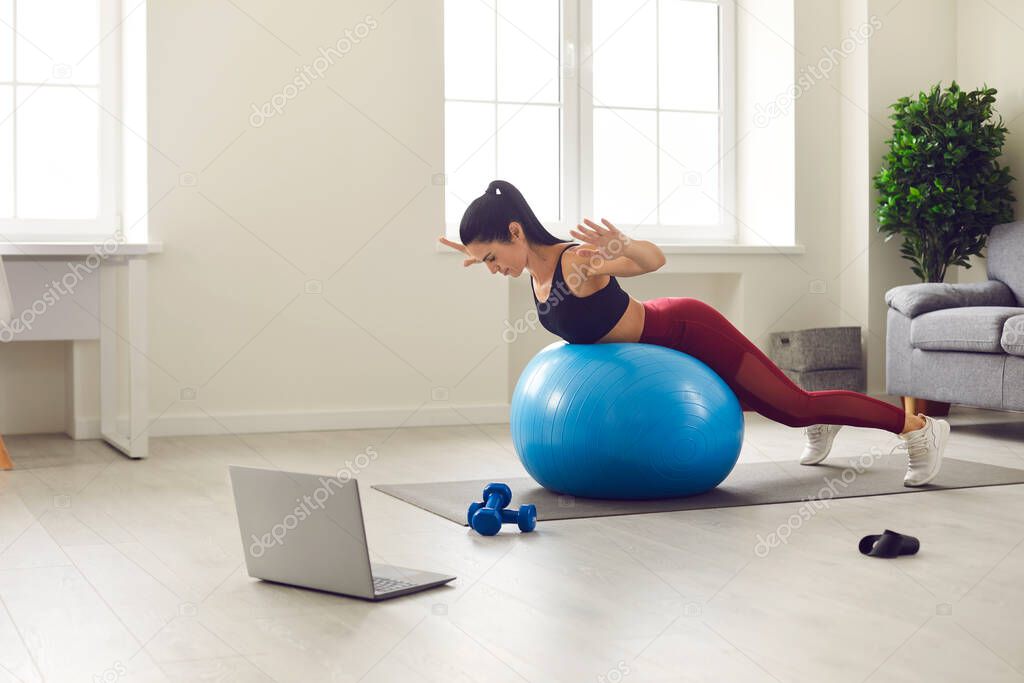 Athlete trains at home in the morning with a fitness ball and watches her favorite show on a laptop.