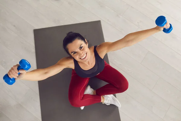 Mujer deportiva sentada en una colchoneta de gimnasio, haciendo ejercicio con pesas y sonriendo a la cámara — Foto de Stock