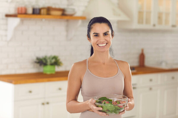 Portrait of slim woman in sports top holding bowl of vegetable salad and smiling at camera