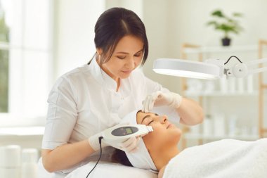 Gülümseyen dermatolog, genç bir kadın için ultrason cihazı yüz temizliği yapıyor.