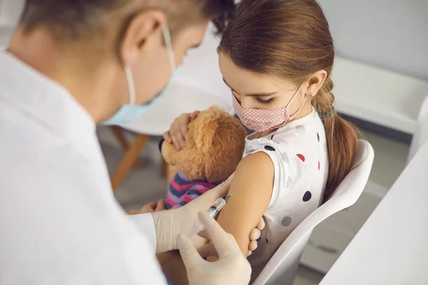 Liten flicka i skyddande medicinsk ansiktsmask sitter med leksak och få vaccination — Stockfoto