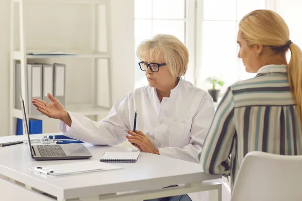 A laptopra mutató idősebb női orvos elmondja a betegnek a kezelés részleteit.. — Stock Fotó