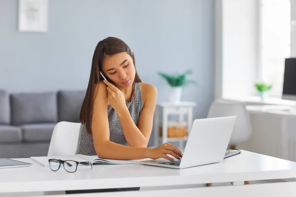 Ernsthaft beschäftigte junge Frau telefoniert und tippt gleichzeitig auf Laptop-Tastatur — Stockfoto