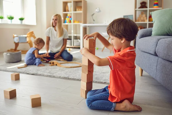 Netter kleiner Junge baut Holzblockturm mit kleinem Bruder und Mama im Hintergrund — Stockfoto