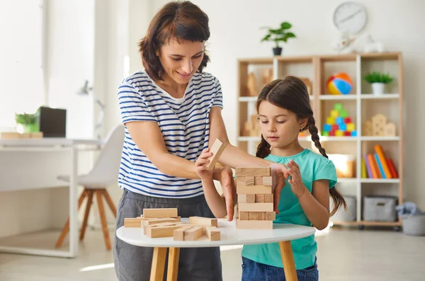 Madre feliz e hija sonriente sentada en el suelo y mirando la pirámide de madera en casa —  Fotos de Stock