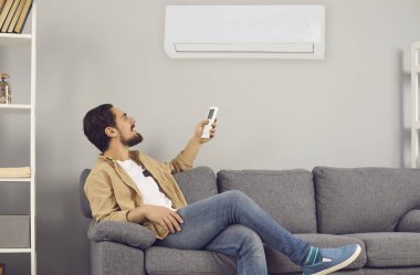 Evdeki kanepede oturan ve uzaktan kumandayla klimayı açan genç adam.