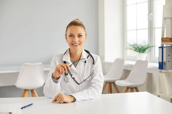 Feliz joven doctora sentada a la mesa en su oficina, sonriendo y mirando a la cámara —  Fotos de Stock