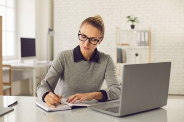 Odaklanmış kadın internet kursuna gidiyor, web sitesi izliyor ve defterine bir şeyler yazıyor.