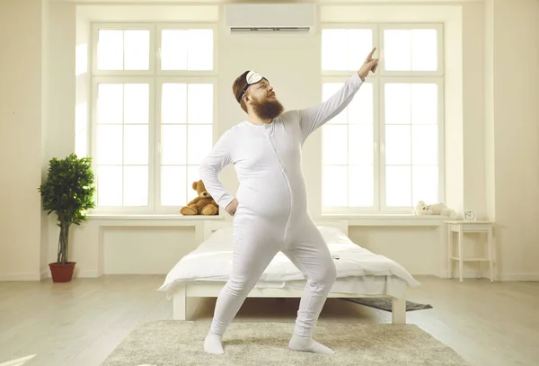 Cheerful chubby young man in pajamas dancing in light sunny bedroom in the morning