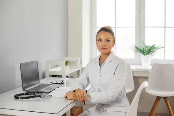 Médecin de famille féminin regardant la caméra alors qu'il était assis à un bureau près d'un ordinateur portable dans son bureau. — Photo