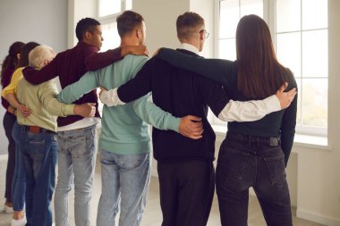 Çeşitli insanlardan oluşan bir grup, sırtı kameraya dönük bir şekilde kucaklaşır ve pencereden dışarı bakar..