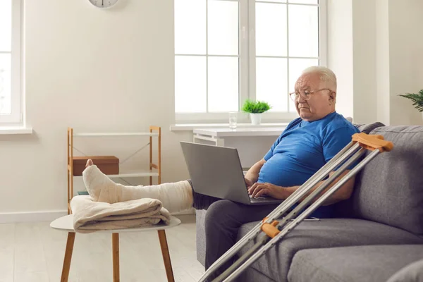 Grave uomo anziano con gamba rotta nel cast seduto sul divano a casa e utilizzando il computer portatile — Foto Stock