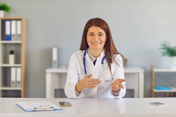 Uśmiechnięta młoda kobieta lekarz siedzi i gestykulacji podczas spotkania online z pacjentem — Zdjęcie stockowe