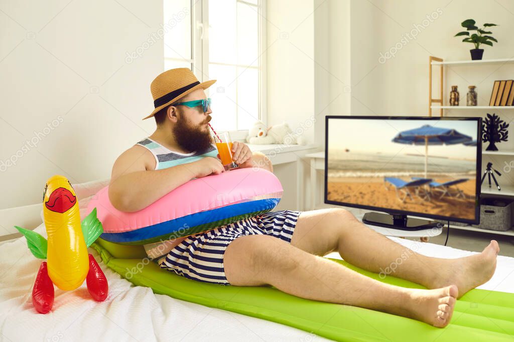 Funny man with inflatable beach toys sipping cocktail and watching travel TV show in lockdown