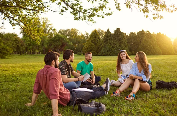 夏の公園で草の上に座ってコミュニケーションをとる若者たち。友人は彼らの自由な時間でリラックス. — ストック写真