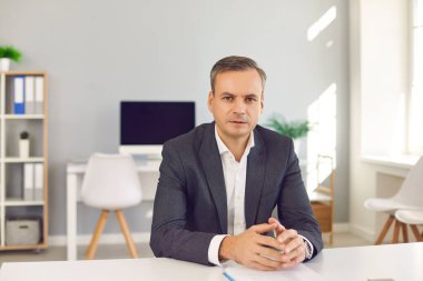 Ofis masasında oturmuş, kameraya bakıyor ve seni dinliyor.