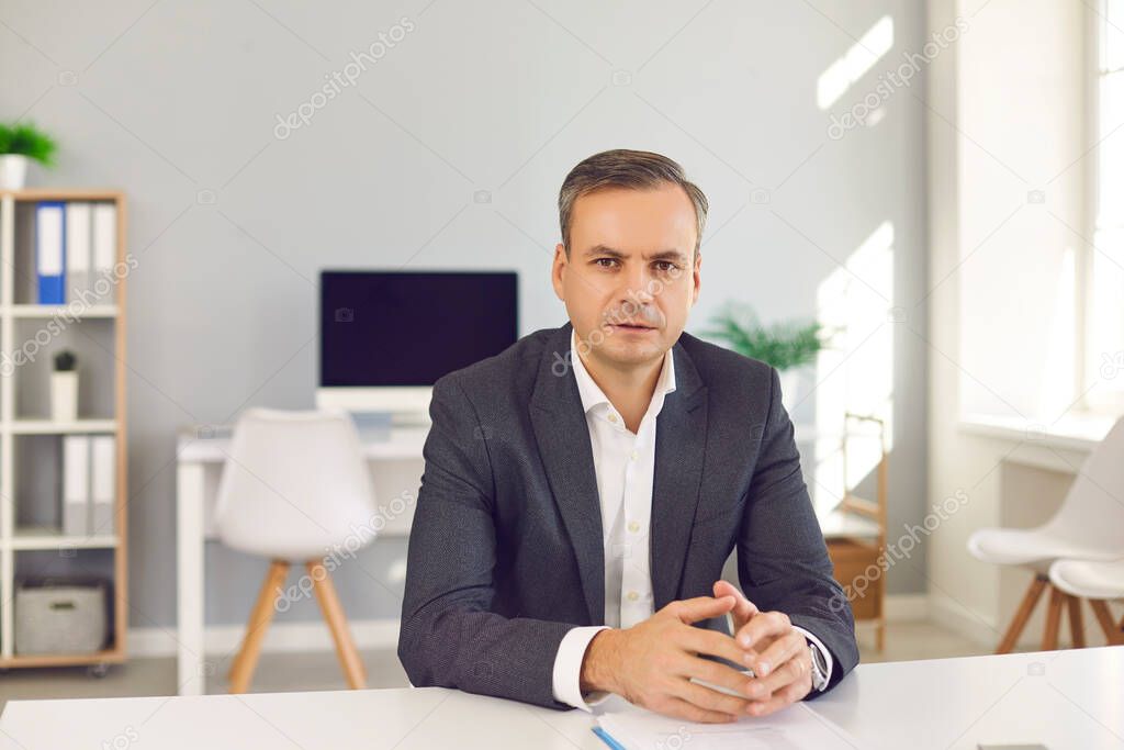 Serious confident businessman sitting at office desk, looking at camera and listening to you