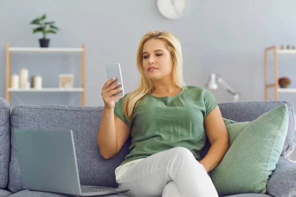 Woman working from home using laptop computer while reading text message on mobile phone.