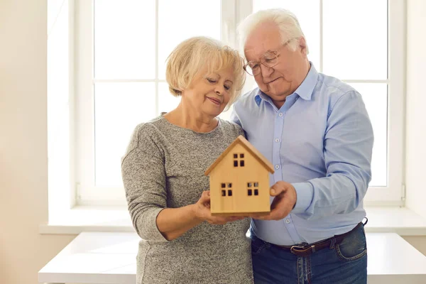 Pareja mayor sonriente mirando un modelo de madera de una casa en sus manos. — Foto de Stock