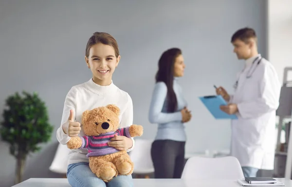 Glückliches kleines Mädchen sitzt in der Arztpraxis, hält Teddy, lächelt und gibt Daumen hoch — Stockfoto