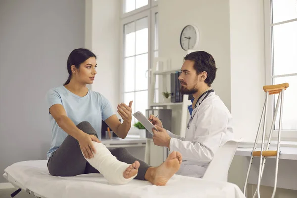 Junge Frau mit Beinbruch spricht während ärztlicher Untersuchung in der Klinik mit Arzt — Stockfoto