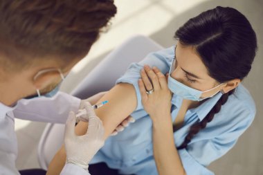 Koruyucu maskeli kadın aşı yapan doktorlara bakıyor.